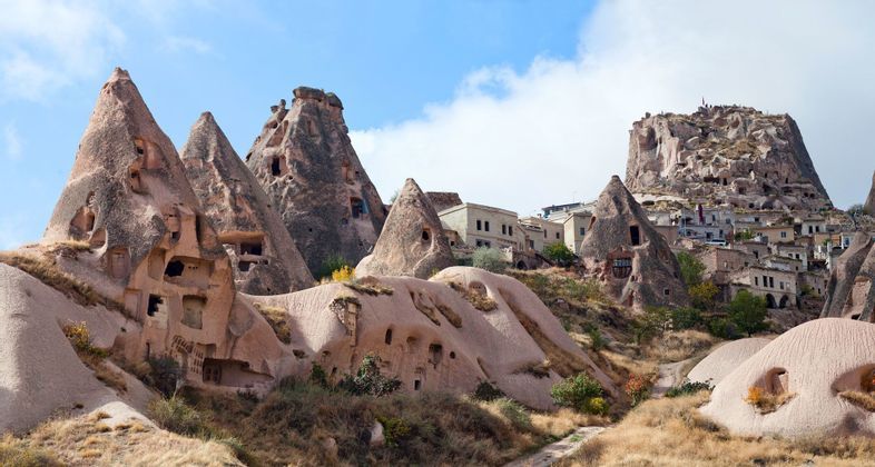 Un villaggio di abitazioni rupestri scavate in formazioni rocciose coniche su un fianco di collina, sotto un cielo parzialmente nuvoloso.
