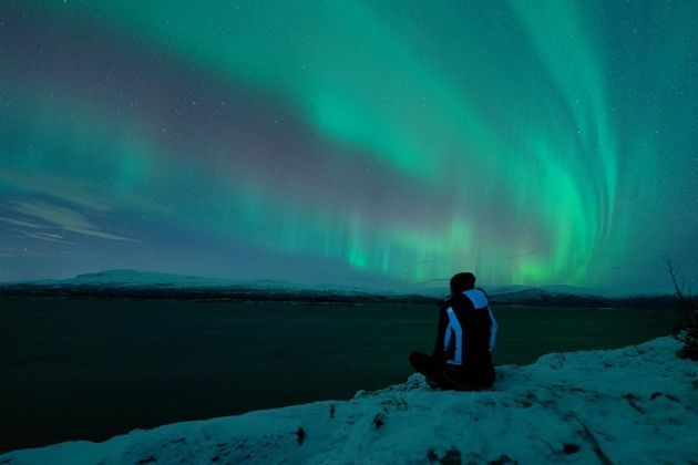 A person sits on a snowy cliff watching the green aurora borealis fill the starry night sky above a dark body of water.