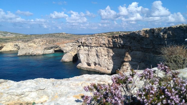 Scogliere rocciose con una formazione ad arco naturale si affacciano su una baia di acqua turchese sotto un cielo blu con nuvole bianche.