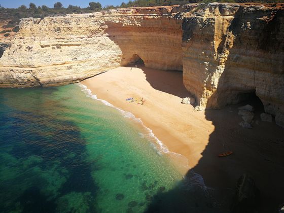 Una playa de arena aislada con aguas cristalinas color turquesa, rodeada de altos acantilados con cuevas, vista desde un ángulo elevado.