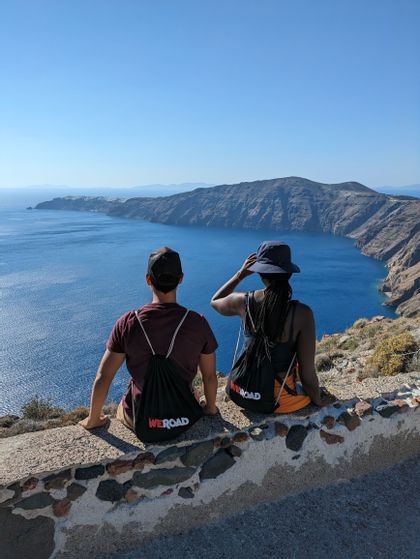 Two people from a WeRoad group trip sit on a stone wall overlooking a large bay with steep, rocky cliffs under a clear blue sky.