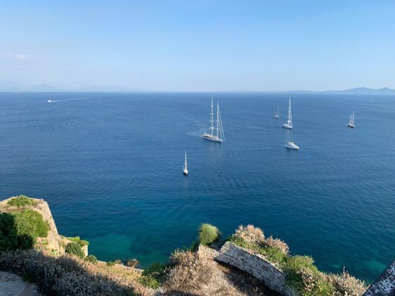 Vogelperspektive von einer felsigen Klippe auf mehrere Segelboote und Yachten auf einem weiten, tiefblauen Meer unter klarem Himmel.