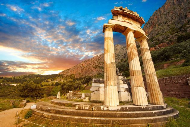 Le rovine di un antico tempio circolare con colonne sorgono su una base di pietra in una verde valle montuosa al tramonto.