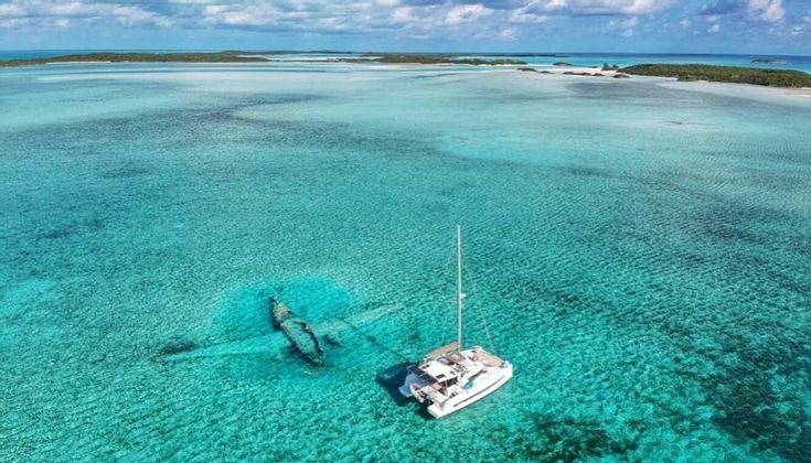 Una vista aerea di un catamarano bianco ancorato vicino a un relitto sommerso in acque basse, limpide e turchesi, con isole all'orizzonte.