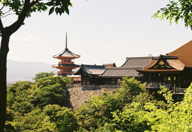 Una pagoda tradicional japonesa y un complejo de templos encaramados en un muro de piedra sobre exuberantes árboles verdes, con montañas en la distancia.