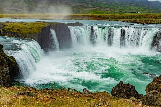 Una cascata ampia e potente precipita da scogliere muschiose in un fiume turbolento color turchese sotto un cielo nuvoloso.