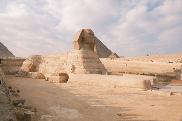 La Grande Sfinge si erge in un paesaggio sabbioso, con una grande piramide visibile sullo sfondo sotto un cielo parzialmente nuvoloso.
