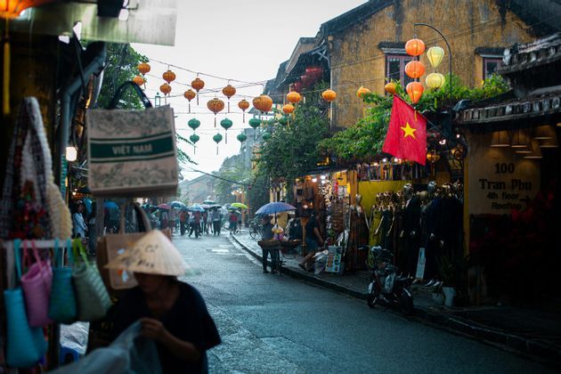 Menschen mit Regenschirmen spazieren eine regnerische vietnamesische Straße entlang, die von Geschäften gesäumt und mit bunten Papierlaternen geschmückt ist.