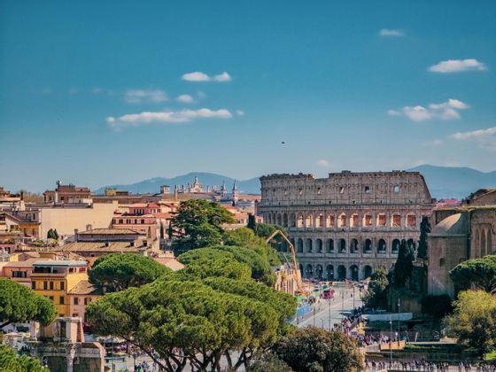 Un paesaggio urbano diurno con il Colosseo, circondato da edifici storici e alberi verdi lussureggianti sotto un cielo azzurro.