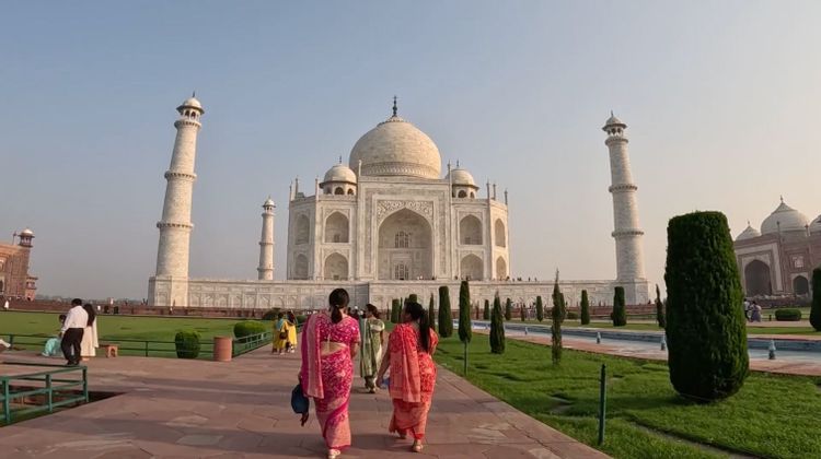 Two women in colorful saris walk on a stone pathway towards a large, white marble mausoleum with manicured gardens.
