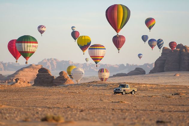 Mongolfiere colorate fluttuano su un paesaggio desertico con formazioni rocciose e un pickup sulla sabbia.