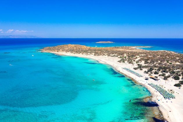 Vista aérea de una península arenosa con playa blanca, agua turquesa y filas de sombrillas bajo un cielo azul claro.