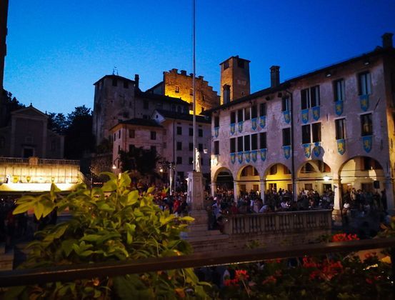 Una folla si raduna in una storica piazza al tramonto, con edifici medievali illuminati e un castello su una collina sullo sfondo.