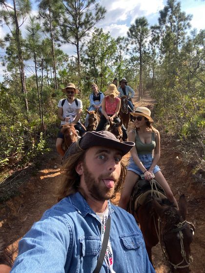 A man takes a selfie, sticking his tongue out, as a WeRoad group trip rides horses on a forest trail.