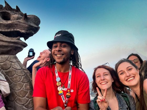 A WeRoad group trip poses for a smiling selfie next to a large stone dragon statue under a blue sky.