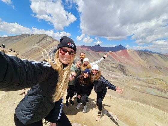 Un viaggio di gruppo WeRoad scatta un selfie sorridente su un crinale montuoso, con catene montuose colorate sullo sfondo sotto un cielo azzurro.