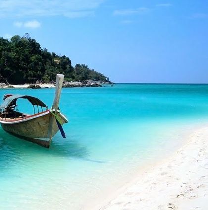 Ein traditionelles Longtail-Boot liegt in ruhigem, türkisfarbenem Wasser vor einem weißen Sandstrand, im Hintergrund eine bewaldete Insel.
