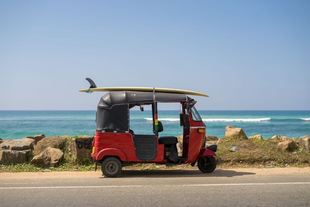 Un tuk-tuk rosso con una tavola da surf legata al tetto è parcheggiato a bordo strada vicino all'oceano sotto un cielo azzurro e limpido.