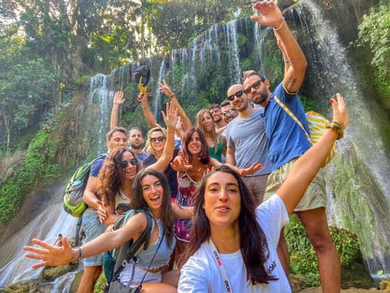 Un voyage en groupe WeRoad de personnes souriantes et saluant pour un selfie devant une cascade entourée d'une végétation luxuriante.