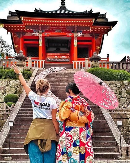 A WeRoad group leader and a woman in a kimono with a pink parasol stand at the base of stairs leading to a red Japanese temple.
