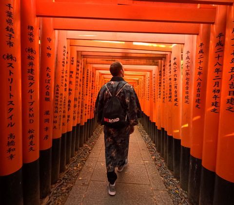 Una persona di spalle che indossa un kimono e uno zaino WeRoad percorre un sentiero fiancheggiato da una lunga serie di torii arancioni.