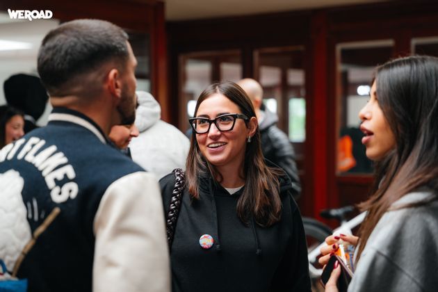 A woman with glasses and a black hoodie smiles while talking with two other people in a WeRoad group.