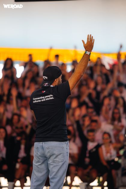 A man, viewed from behind, wears a WeRoad t-shirt and raises his hand to a large, blurred crowd at an event.