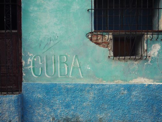 Le mot 'CUBA' est peint sur un mur patiné, turquoise et bleu, à côté d'une fenêtre à barreaux.