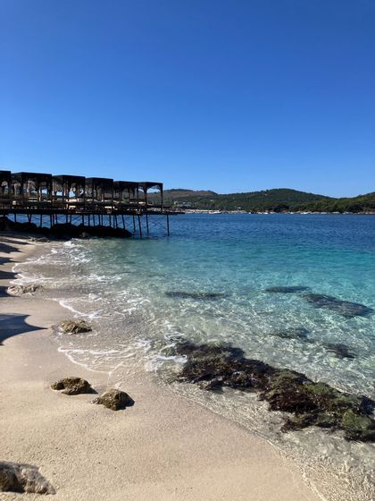 Clear turquoise water gently waves onto a sandy shore next to a wooden pier with cabanas on a sunny day.