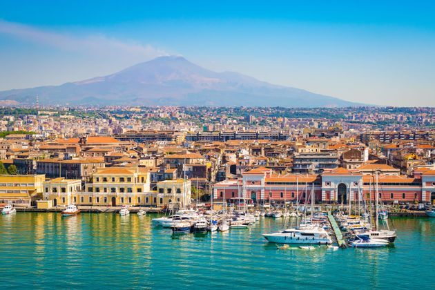 Un porto di città costiera, colmo di barche, con una grande montagna che si erge sullo sfondo sotto un cielo azzurro e limpido.