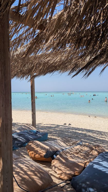 Des poufs sous un auvent de chaume donnent sur des gens qui nagent dans les eaux turquoise et transparentes de la plage.