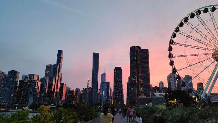 People strolling along the promenade next to a massive Ferris wheel and modern city skyscrapers, all under a pink sunset sky.
