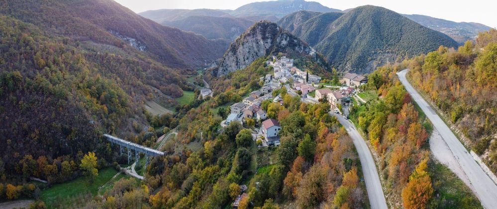 Una vista aerea di un villaggio incastonato tra le montagne con alberi autunnali colorati, una strada tortuosa e un ponte in una valle.