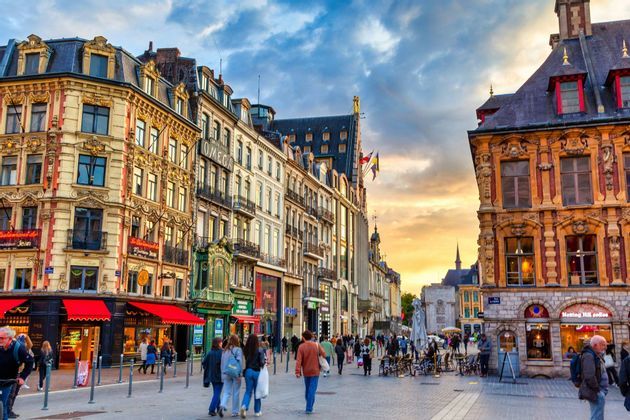 Una piazza del centro di Lille al tramonto, con le facciate fiamminghe, i negozi illuminati e la gente a passeggio.