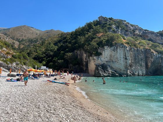 Des personnes se détendent et nagent le long d'une plage de galets nichée à côté de falaises rocheuses abruptes et d'une eau turquoise sous un ciel bleu.