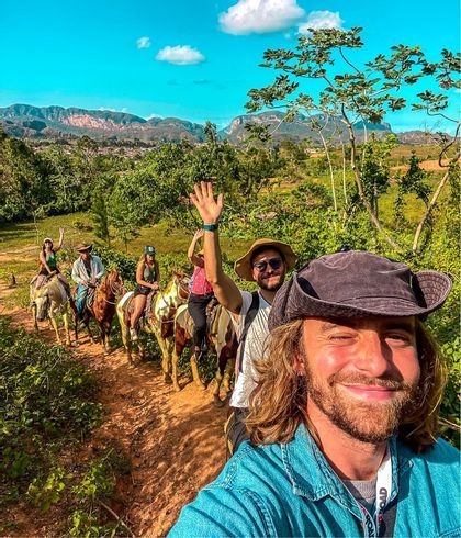 A man takes a selfie of his WeRoad group trip horseback riding on a trail through a lush green valley with mountains in the background.