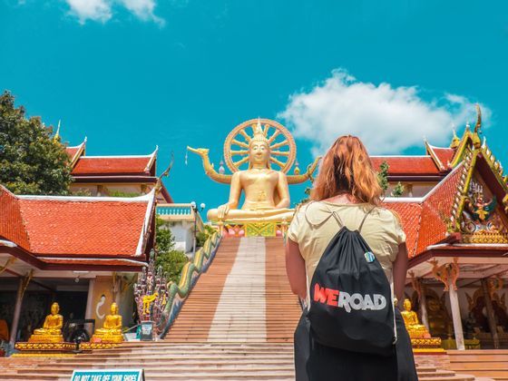 A woman with a WeRoad backpack stands at the bottom of a staircase looking up at a giant golden Buddha statue.