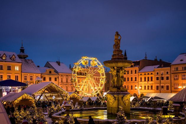 Una ruota panoramica illuminata e bancarelle festive circondano una fontana di pietra illuminata in una piazza cittadina innevata al crepuscolo.