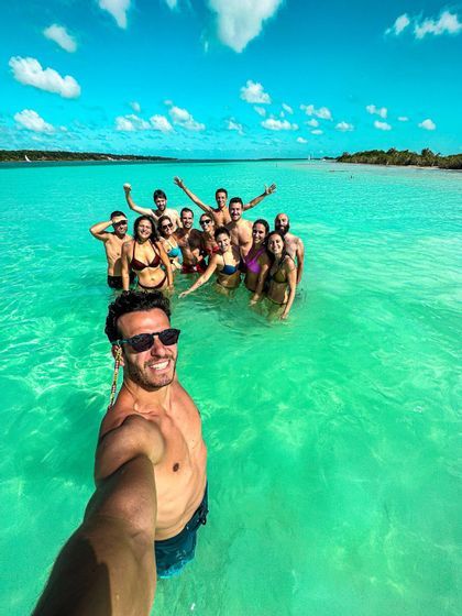 A WeRoad group, all smiles, taking a selfie while standing in clear turquoise water under a bright blue sky.