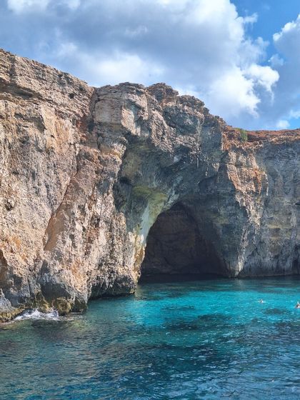 Falaises rocheuses escarpées avec une grande ouverture de grotte naturelle au-dessus d'une eau turquoise claire sous un ciel partiellement nuageux.