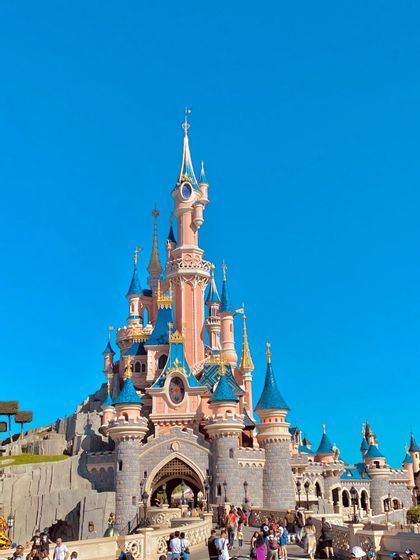 Un castello da fiaba rosa con torri dal tetto blu si erge imponente sotto un cielo azzurro limpido, con folle di persone alla sua base.