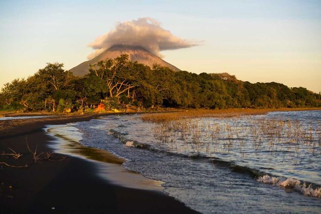 Un vulcano con la cima avvolta da una nuvola si erge dietro una foresta lussureggiante, visto da una spiaggia di sabbia scura con onde che lambiscono la riva.