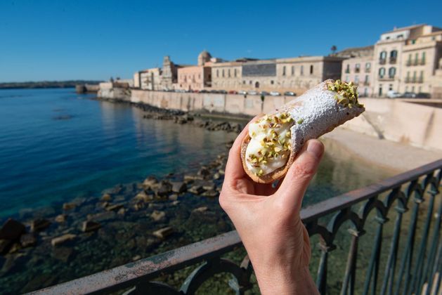 Una mano tiene un cannolo con pistacchi e zucchero a velo davanti a una vista panoramica di una città costiera.