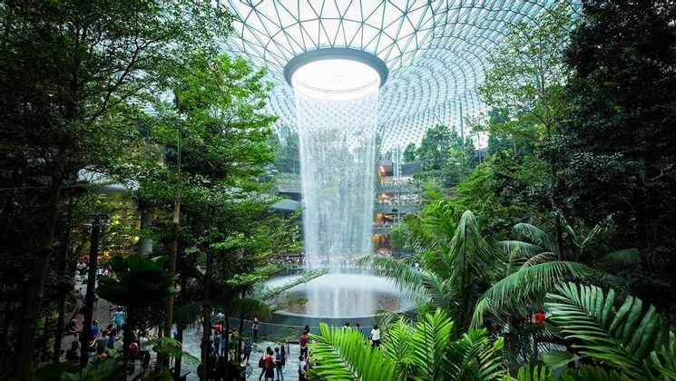 Une grande cascade intérieure dévale d'un plafond en dôme de verre dans un jardin luxuriant et à plusieurs niveaux, rempli d'arbres et de personnes.