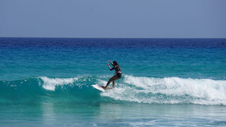Una donna in muta fa surf su una tavola bianca, cavalcando un'onda turchese nell'oceano blu sotto un cielo sereno.