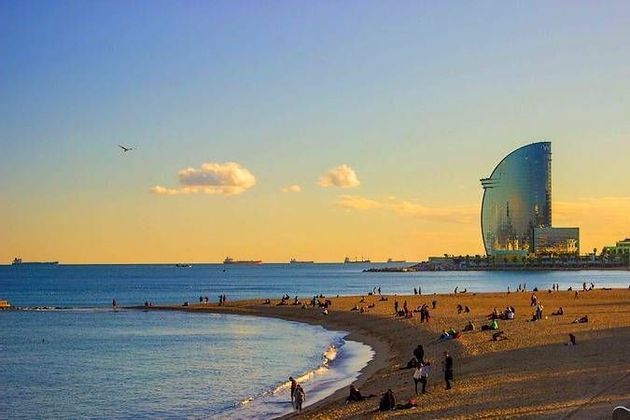 People relaxing on a sandy beach during sunset, with a large, sail-shaped glass building next to the sea in the background.