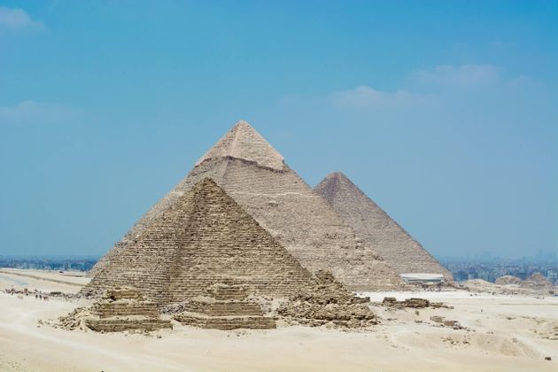 Tres grandes y antiguas pirámides se alzan en un desierto arenoso bajo un cielo azul claro, con ruinas más pequeñas en primer plano.