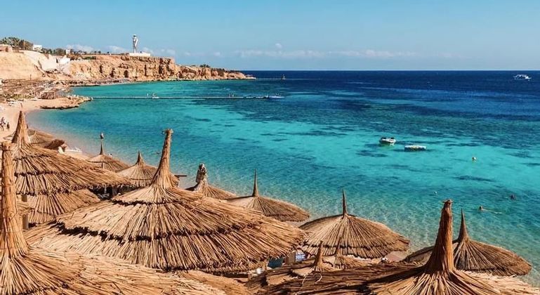 Vista aérea de sombrillas de paja en una playa de arena con vistas a una bahía turquesa y un acantilado rocoso bajo un cielo azul claro.