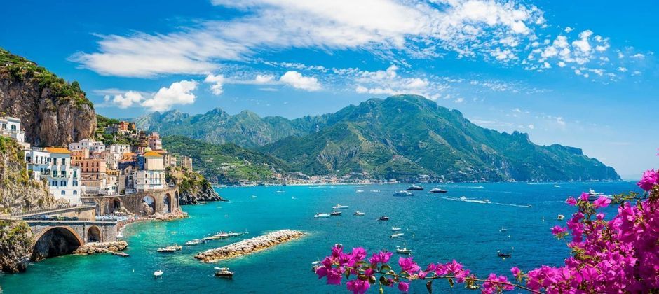 A colorful cliffside town overlooks a turquoise bay filled with boats, with green mountains and a blue, cloudy sky in the background.