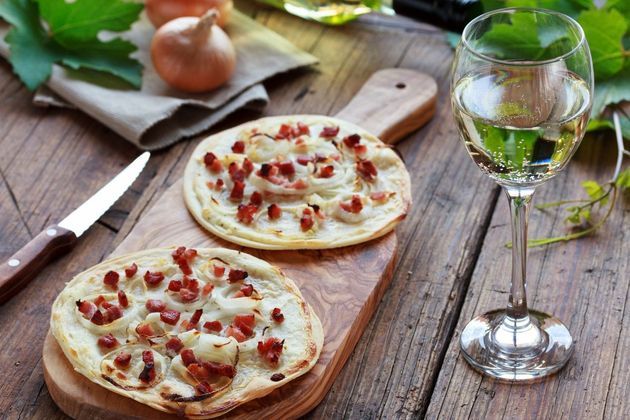 Due Flammkuchen alsaziani con pancetta e cipolle su un tagliere di legno accanto a un bicchiere di vino bianco su un tavolo rustico.